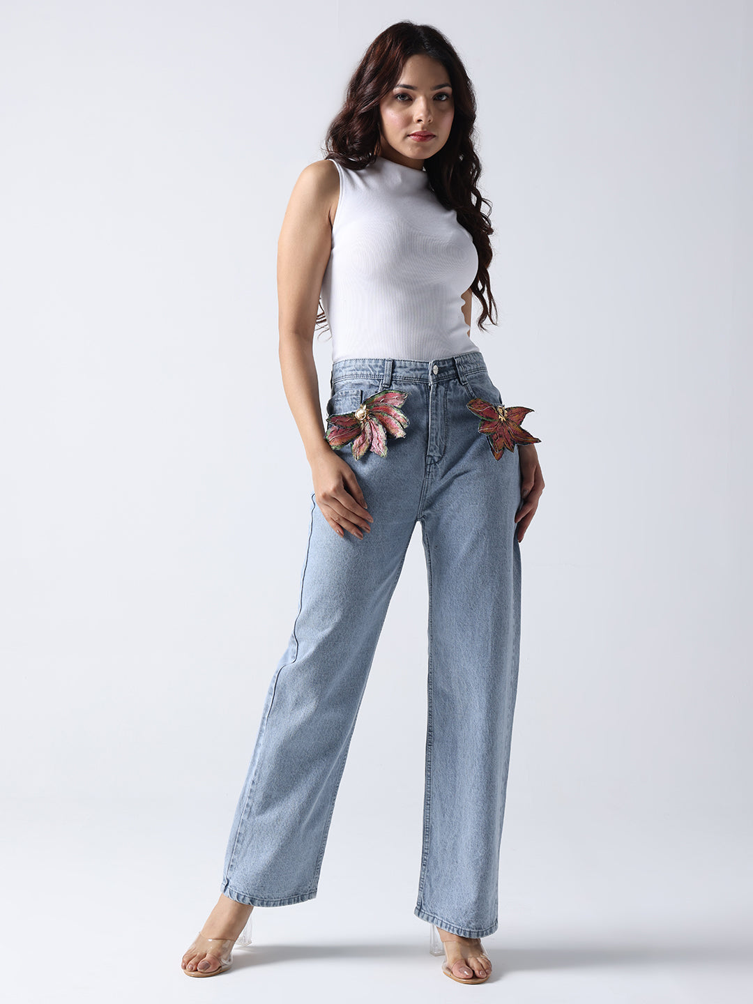 Model wearing light blue jeans with floral brooch on jeans paired with white sleeveless top
