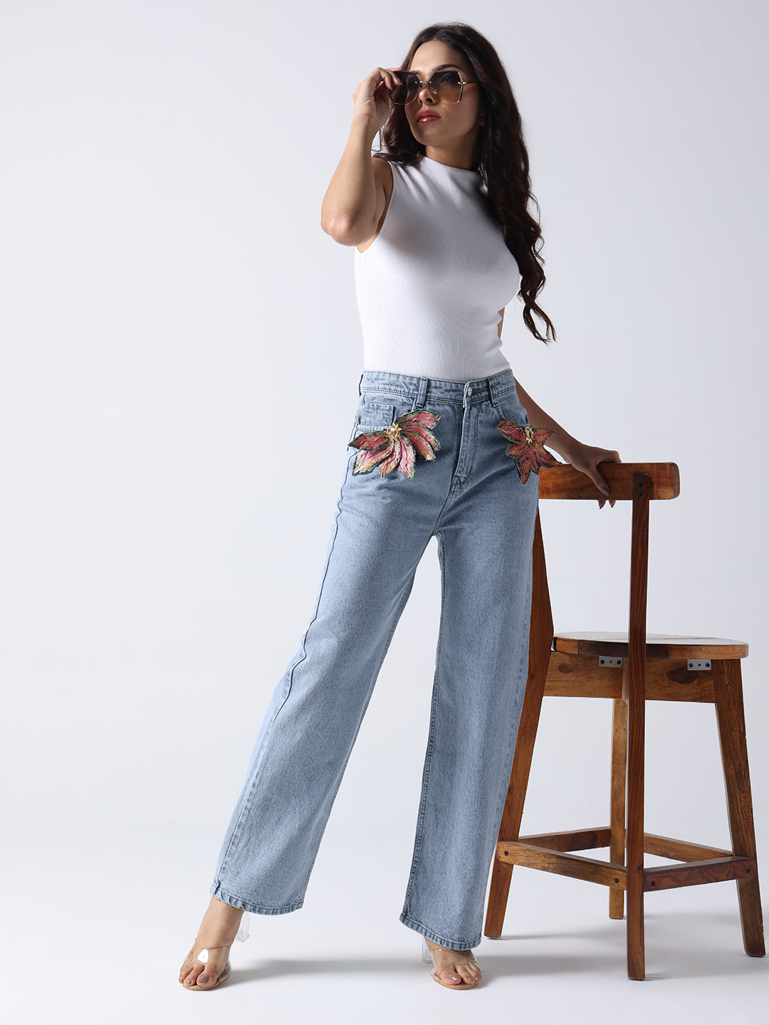 Woman wearing light blue jeans with floral brooch on jeans paired with white sleeveless top and sunglasses