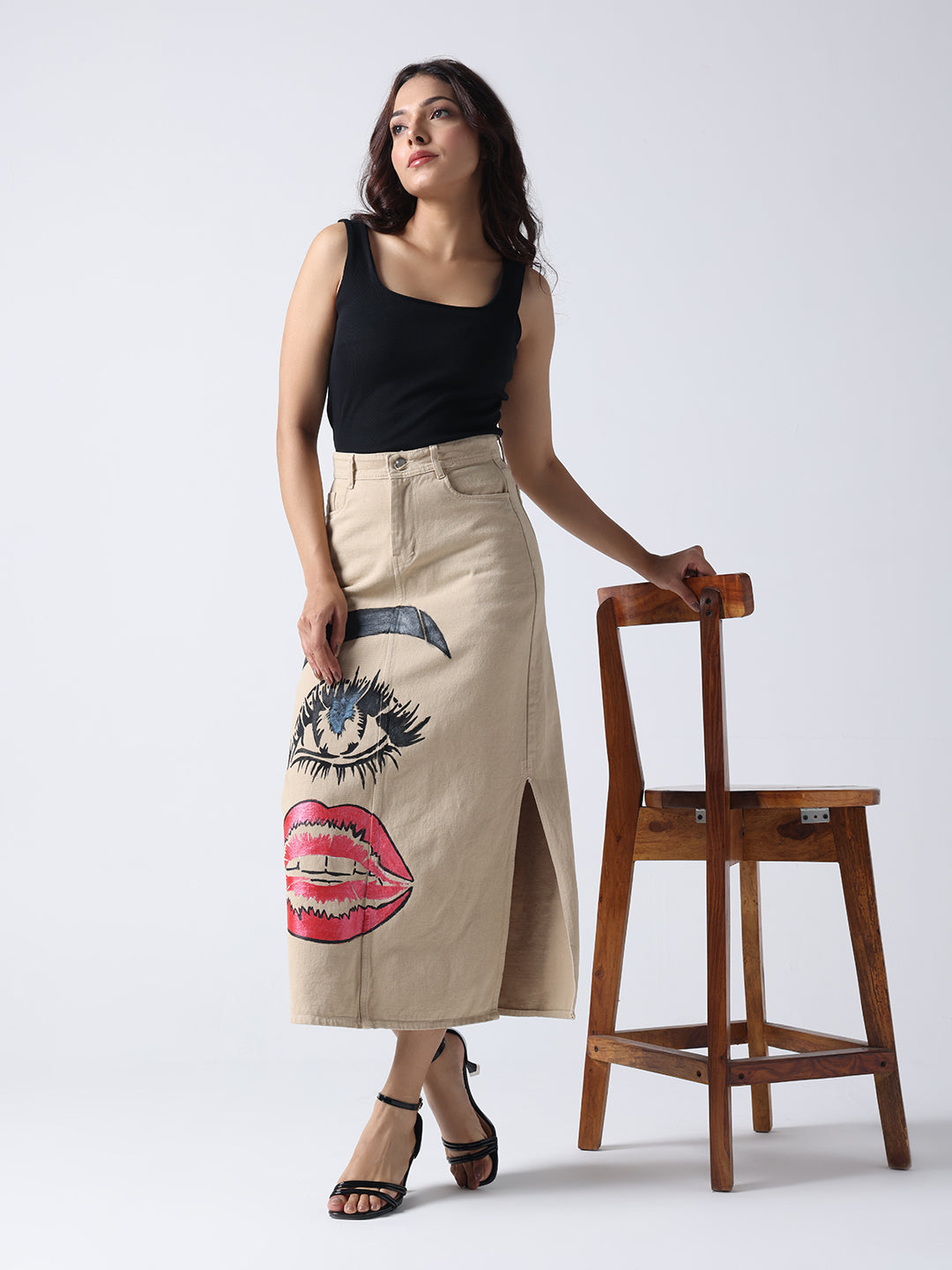Woman wearing Gotamoda Pop Art Glam Hand Painted Denim Skirt with black top and heels standing next to wooden chair
