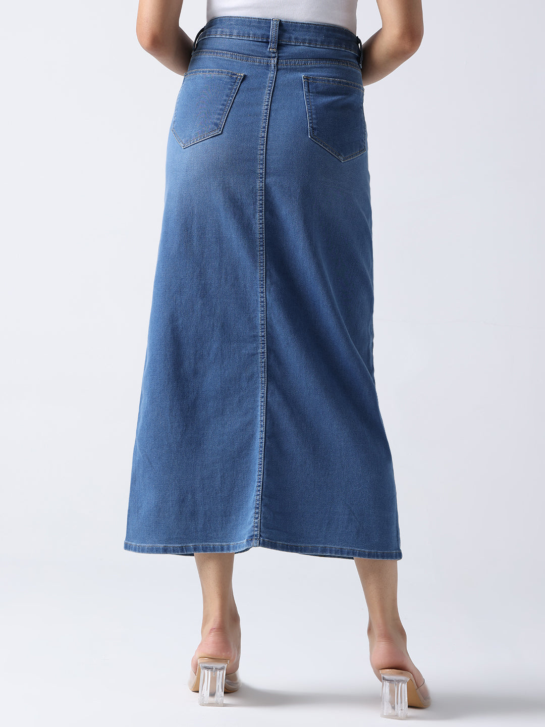 Back view of queen bee denim midi skirt in blue worn by model with white top and clear heels