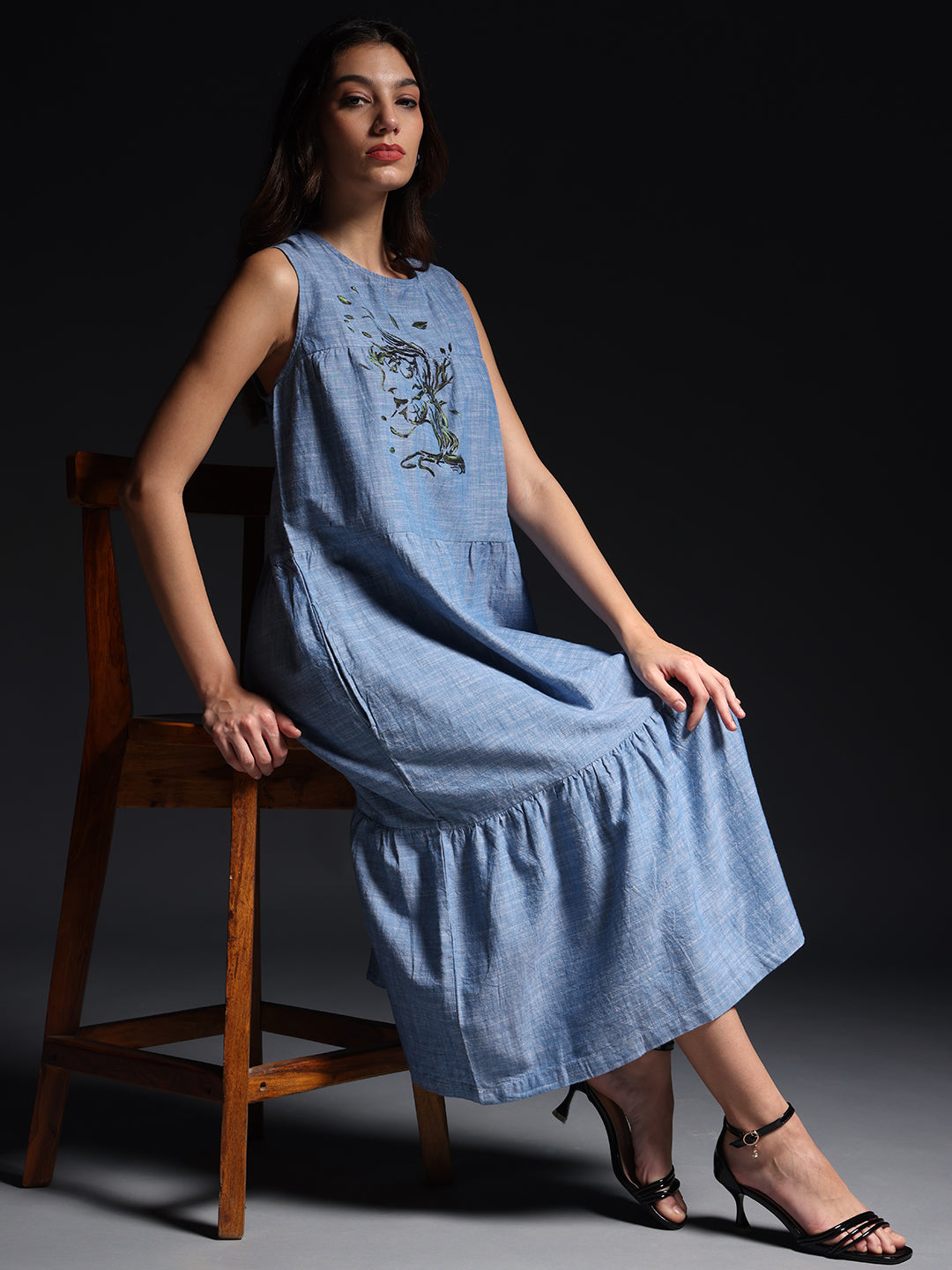 Gotamoda sleeveless maxi dress in light blue with floral embroidery, worn by model sitting on wooden chair
