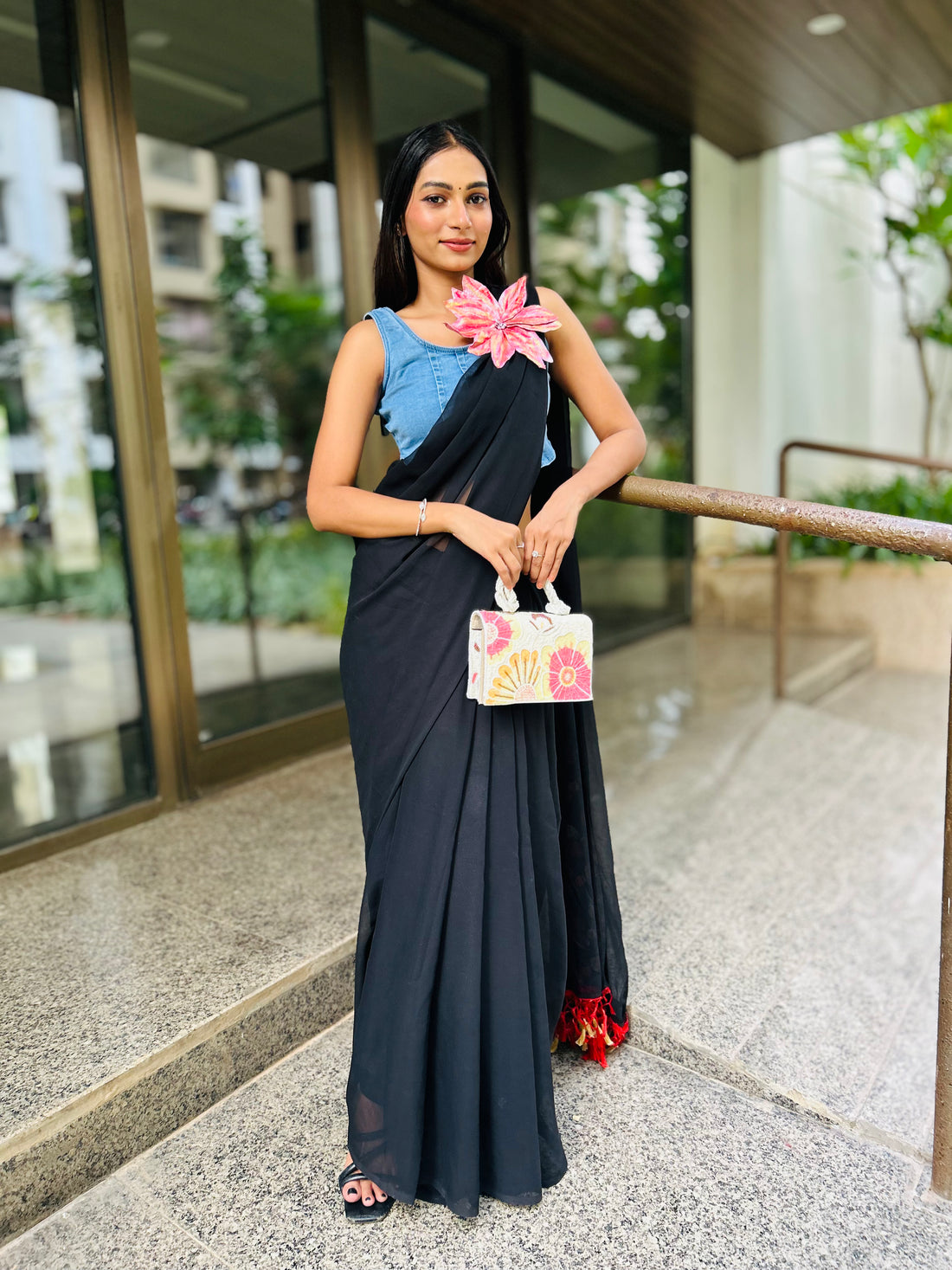 Woman wearing black saree with denim blossom brooch and blue blouse holding floral handbag outdoors