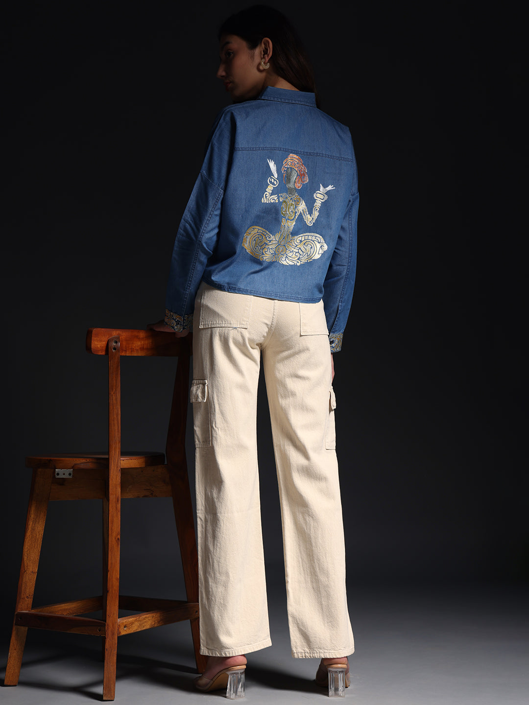 Woman wearing a denim shirt with white jeans standing next to a wooden chair against a dark background