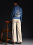 Woman wearing a denim shirt with white jeans standing next to a wooden chair against a dark background
