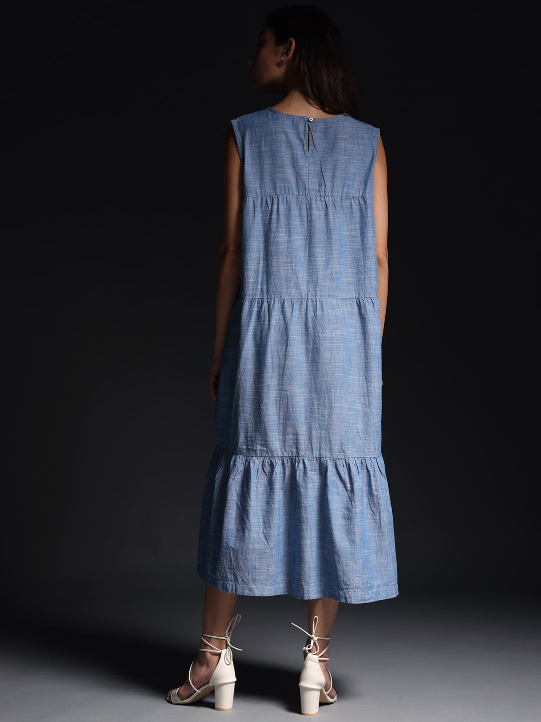 Back view of model wearing Gotamoda blue sleeveless maxi dress with tiered design and white heeled sandals